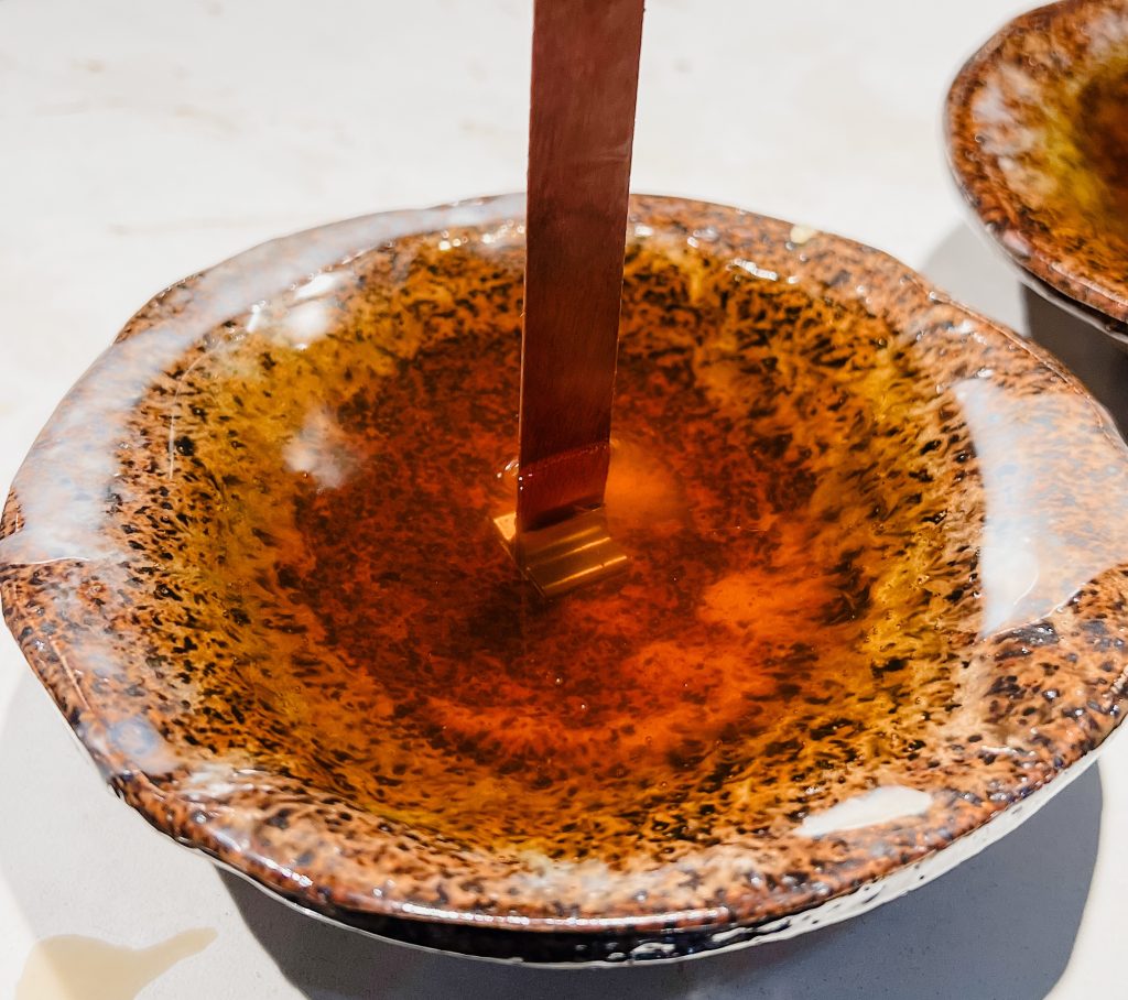 A handmade beeswax candle that had been poured in a handmade ceramic bowl. The beeswax candle has a wood wick.the ceramic bowl is rust colored with black speckles.