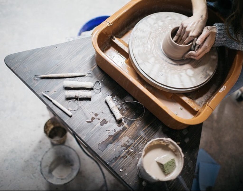 A woman throwing pottery bowl on a potter’s wheel. A ceramic sponge, ceramic loop tool, and ceramic wire cutters are on the pottery table.
