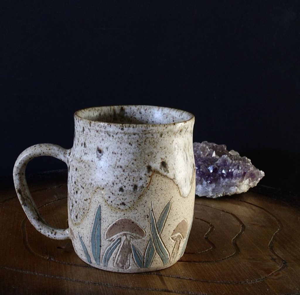 Speckled stoneware mug with underglaze mushrooms and a black and white speckled glaze on a black background