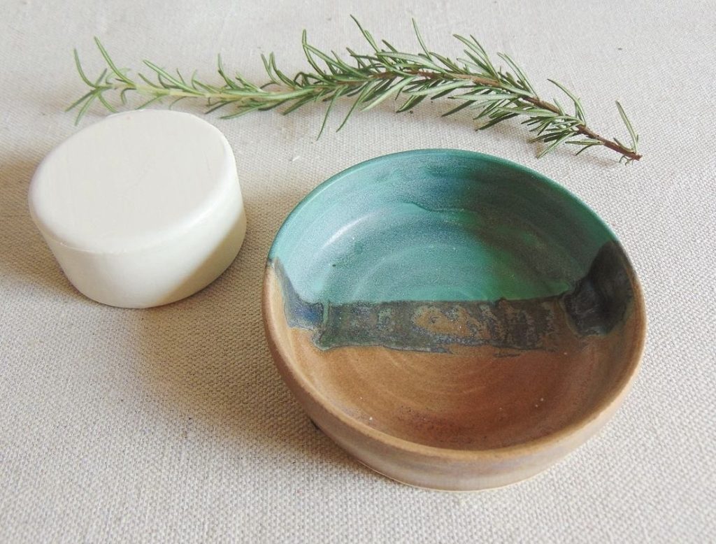Turquoise and gray handmade ceramic soap dish next to a white round bar of soap laying on a white tablecloth