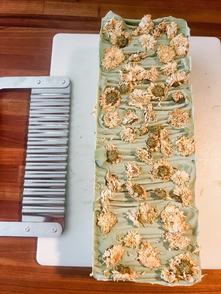 Handmade cold process soap ready to be cut. Cold process soap loaf on wooden countertop with soap cutter next to it. Nontoxic aloe soap with dried flowers on top.