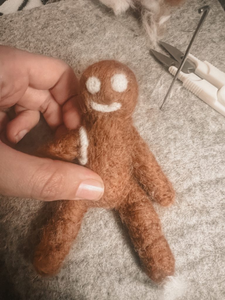 Hand holding a needle felted gingerbread man on a felt pad  with needle felting needle and scissors in the background 