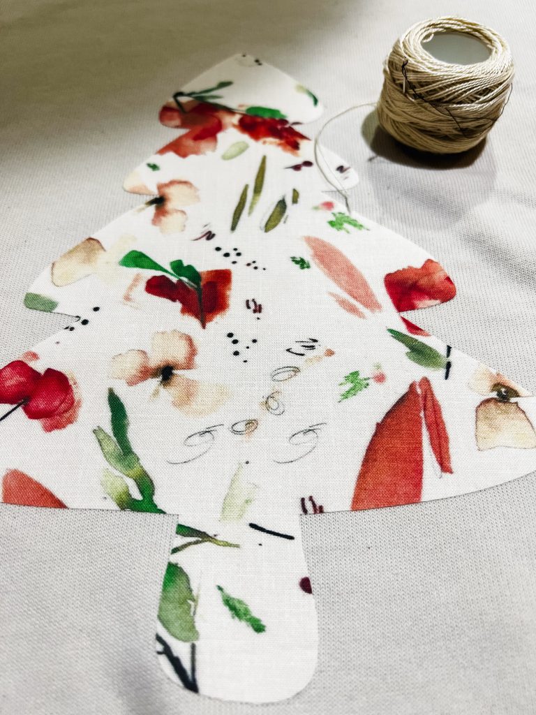 An iron on Christmas tree fabric applique on a sweatshirt made with the Cricut maker 3. A needle and embroidery thread sit next to the sweatshirt.