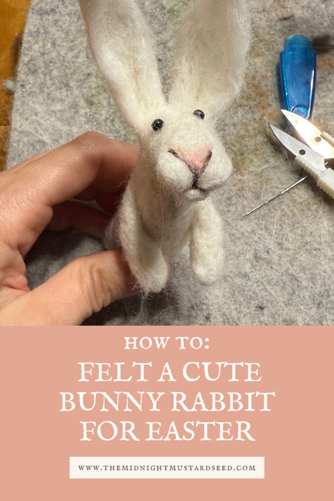 How to felt a cute bunny rabbit for Easter. White needle felted rabbit on a felt pad with a felting needle and needle felt scissors in the background. Needle felting tutorial by the midnight mustard seed