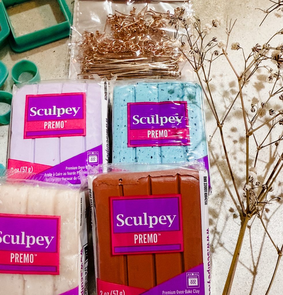 A photo showing materials needed to make polymer clay earrings. This photo shows terracotta and turquoise sculpey premo clay, clay earring cutters, and earring hooks on a white surface.