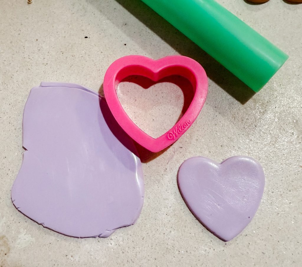 A lavender slab of polymer clay sculpey premo with a heart cookie cutter and a clay roller. Purple polymer clay heart