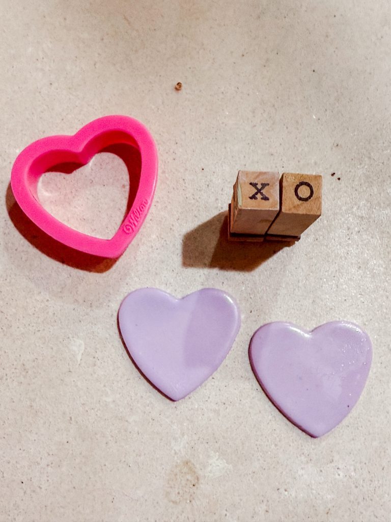 Two purple polymer clay hearts, a heart cookie cutter, and two letter stamps “x” and “o” . How to make valentines heart earrings