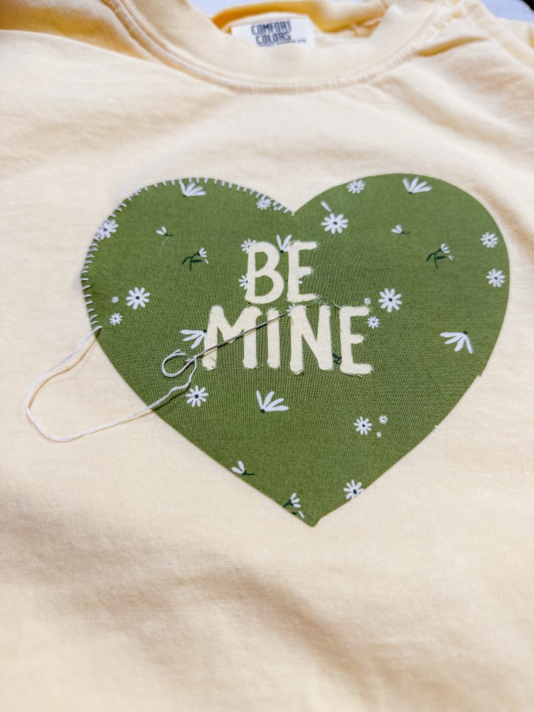 A green valentine heart applique that says ‘be mine’ is being appliquéd into a yellow t shirt using a whip stitch . The midnight mustard seed applique tutorials