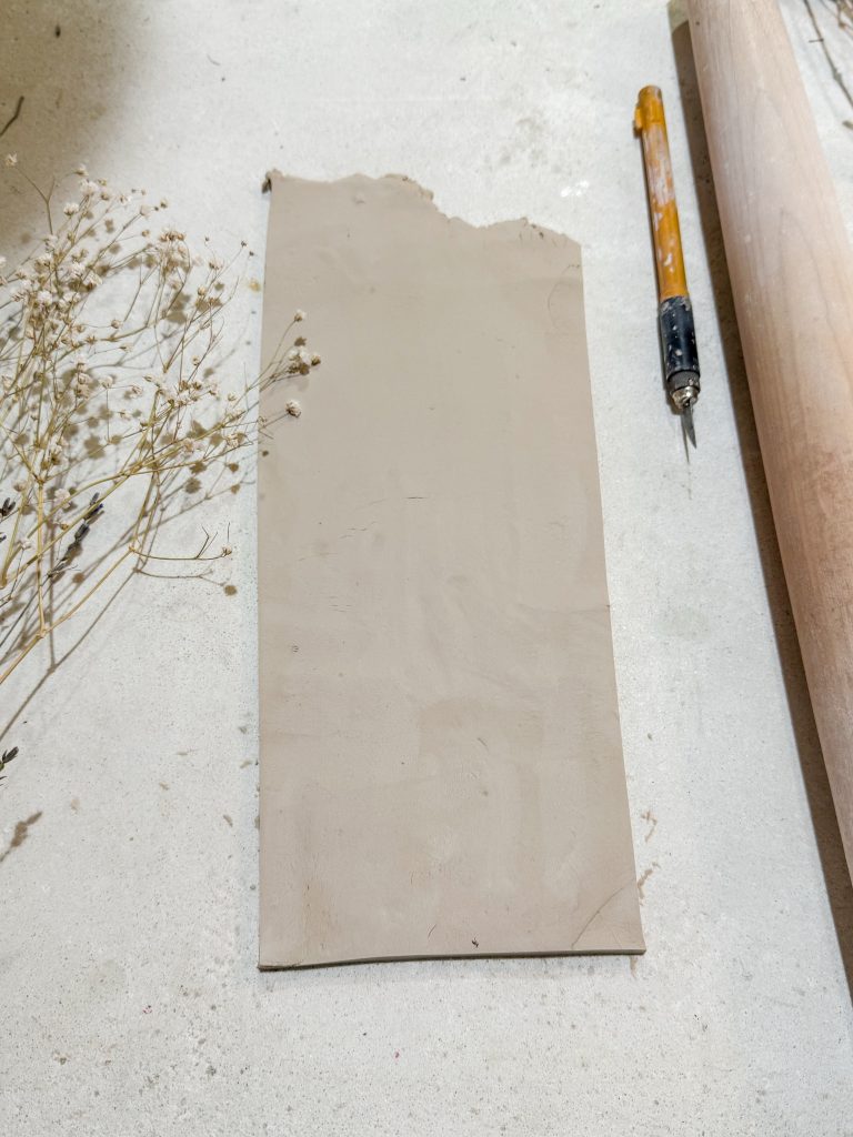 A slab of white pottery clay m, an x acto knife, and a rolling pin on a white concrete work surface. How to make a pottery wall planter by the midnight mustard seed