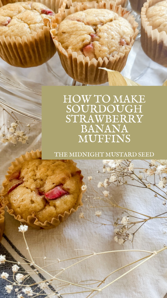 A collection of freshly baked sourdough strawberry banana muffins in brown paper liners, displayed on a glass stand and scattered on a light-colored tablecloth. The muffins are golden brown with visible chunks of strawberries. In the center of the image, a sage green rectangular overlay contains white text that reads, “HOW TO MAKE SOURDOUGH STRAWBERRY BANANA MUFFINS,” with “THE MIDNIGHT MUSTARD SEED” written below in smaller font. Delicate dried baby’s breath flowers are sprinkled around the muffins, adding a rustic touch to the scene. This image is from a recipe blog post on www.themidnightmustardseed.com.