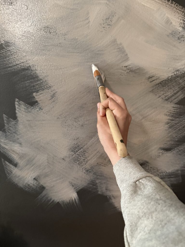 A close-up image of a hand holding a paintbrush, applying white paint in broad, sweeping strokes onto a dark surface. The hand is wearing a gray sleeve, and the paint creates a textured, cloud-like pattern against the darker background.