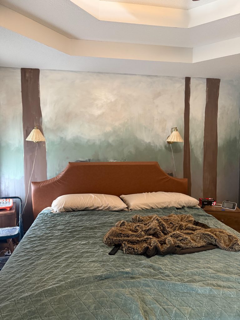 A bedroom with a cozy setup, featuring a bed with a brown leather headboard and white pillows, covered with a teal quilted comforter and a furry brown throw blanket. The wall behind the bed has a partially painted mural of tall trees in shades of brown and green, with a soft gradient background. Two woven lampshades are mounted on the wall, casting a warm glow. On the left, a blue step ladder and a red paint tray with brushes are visible, while on the right, a wooden nightstand holds a small tablet, a box of tissues, and other small items. The ceiling has a modern tray design with recessed lighting.