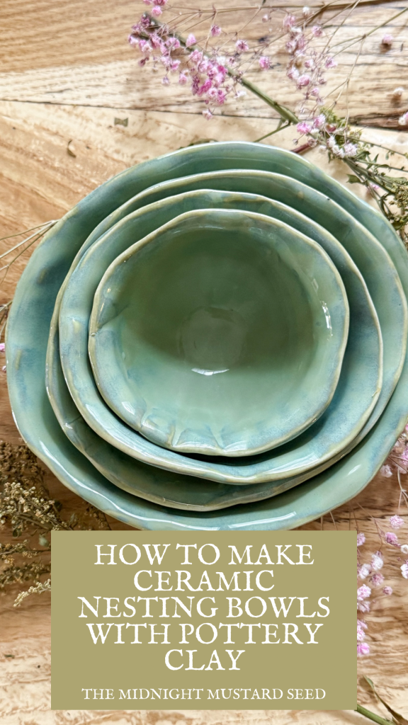 A stack of four handcrafted ceramic nesting bowls in shades of teal and green sits on a rustic wooden surface. The bowls are neatly nested, showcasing their smooth, glossy finish and organic shapes. Delicate pink dried flowers are arranged around the bowls, adding a soft, natural touch to the scene. At the bottom of the image, a text overlay in a mustard yellow box reads, “HOW TO MAKE CERAMIC NESTING BOWLS WITH POTTERY CLAY” in bold white letters, followed by “THE MIDNIGHT MUSTARD SEED” in smaller white text, indicating the blog name.