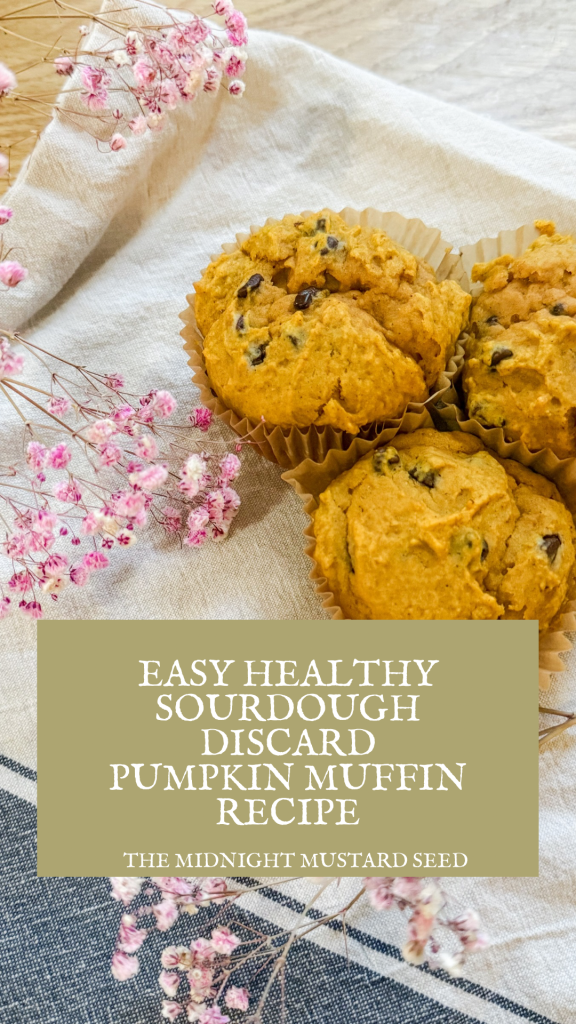 A cozy fall scene featuring three healthy sourdough discard pumpkin muffins with mini chocolate chips, nestled in muffin liners on a rustic white cloth. The muffins are surrounded by delicate pink dried flowers, adding a touch of elegance. A green text box overlay reads ‘EASY HEALTHY SOURDOUGH DISCARD PUMPKIN MUFFIN RECIPE’ in white, with ‘THE MIDNIGHT MUSTARD SEED’ written below, set against a textured blue and white striped fabric background