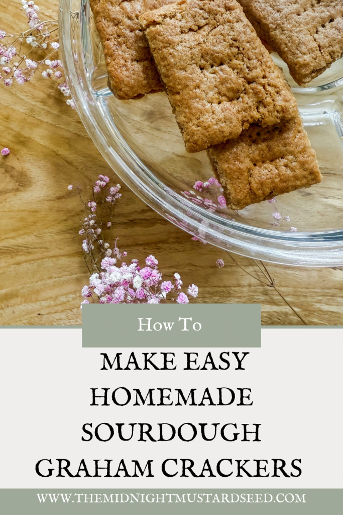 A glass bowl on a wooden surface contains several homemade sourdough graham crackers stacked together. The crackers have a rustic, textured appearance with a golden-brown color. Surrounding the bowl are delicate pink and white dried flowers, adding a decorative touch. The lower part of the image features a text overlay with the title “How To MAKE EASY HOMEMADE SOURDOUGH GRAHAM CRACKERS” in bold black letters on a light background, with a website URL “WWW.THEMIDNIGHTMUSTARDSEED.COM” in a green banner below.