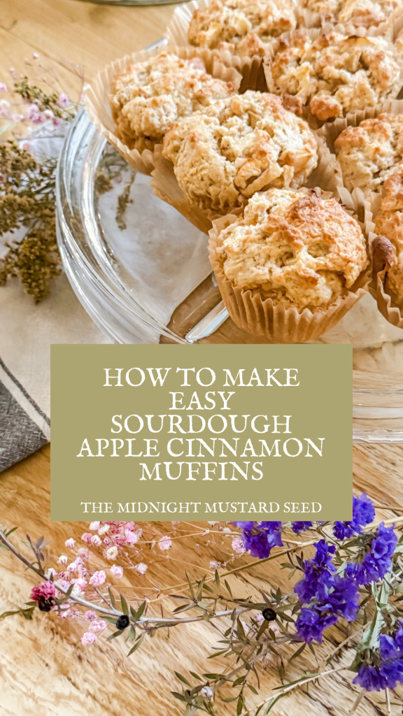 A beautifully arranged platter of freshly baked sourdough apple cinnamon muffins, displayed on a wooden table. The muffins are golden brown with a crumbly topping, placed in a glass dish lined with paper liners. Surrounding the platter are delicate dried flowers in shades of pink and purple, adding a rustic and charming touch. A textured fabric napkin in gray and white is partially visible on the side. The image includes a text overlay that reads, “HOW TO MAKE EASY SOURDOUGH APPLE CINNAMON MUFFINS” with “THE MIDNIGHT MUSTARD SEED” written below, indicating the blog name.