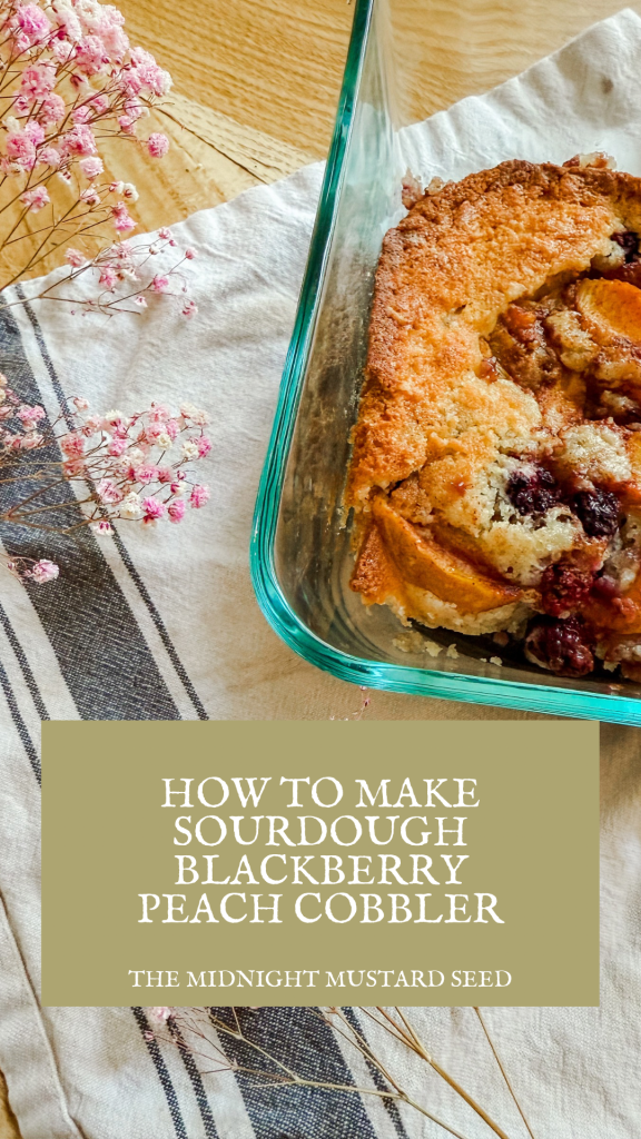 A beautifully styled photo of a freshly baked sourdough blackberry peach cobbler in a glass baking dish, featuring a golden brown biscuit topping with juicy peaches and tart blackberries peeking through. The dish is placed on a wooden table adorned with a white cloth, a striped towel, and delicate pink dried flowers, creating a cozy and rustic atmosphere. A text overlay reads “HOW TO MAKE SOURDOUGH BLACKBERRY PEACH COBBLER” with “THE MIDNIGHT MUSTARD SEED” written below in elegant white font on a green background.