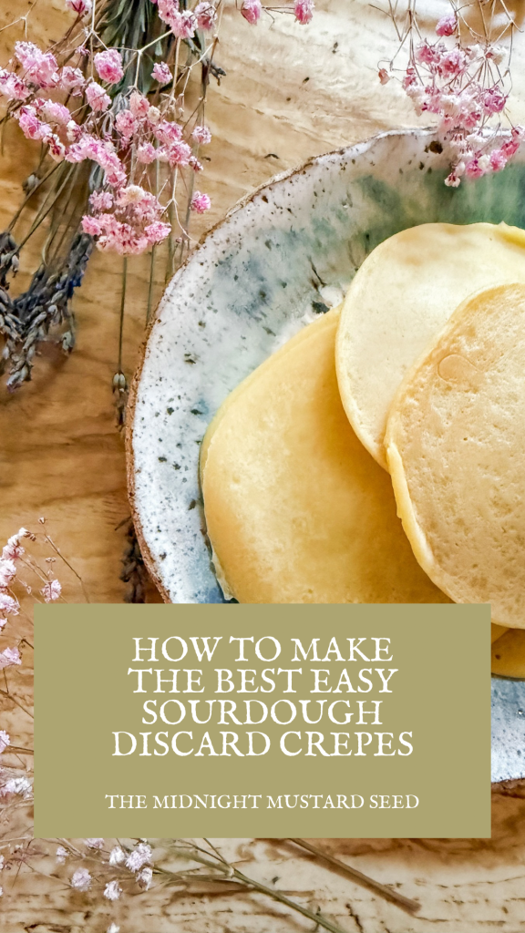 A rustic wooden table features a ceramic bowl filled with golden sourdough discard crepes, their soft, fluffy texture visible. Delicate pink dried flowers and lavender sprigs are artfully arranged around the bowl, adding a touch of natural elegance. A green text box at the bottom of the image reads, “HOW TO MAKE THE BEST EASY SOURDOUGH DISCARD CREPES” in bold white letters, with “THE MIDNIGHT MUSTARD SEED” written below in smaller white text, identifying the blog.