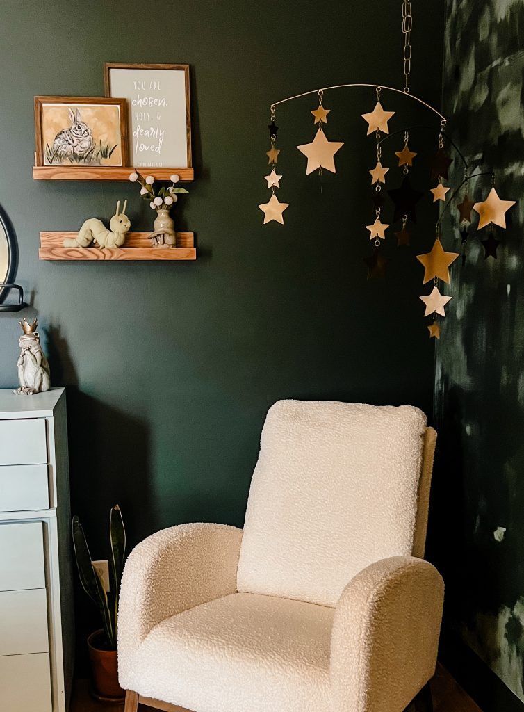 A cozy nursery corner from The Midnight Mustard Seed featuring a plush cream-colored armchair against a dark green wall with abstract texture. Above, a wooden shelf holds a framed illustration of a rabbit, a sign with the text “you are chosen, holy, & dearly loved” (Colossians 3:12), a small vase with dried flowers, a plush caterpillar, and a frog figurine with a crown. A decorative mobile with gold and black stars hangs from the ceiling, adding a whimsical touch. A potted plant and a white dresser with drawers complete the serene setup.