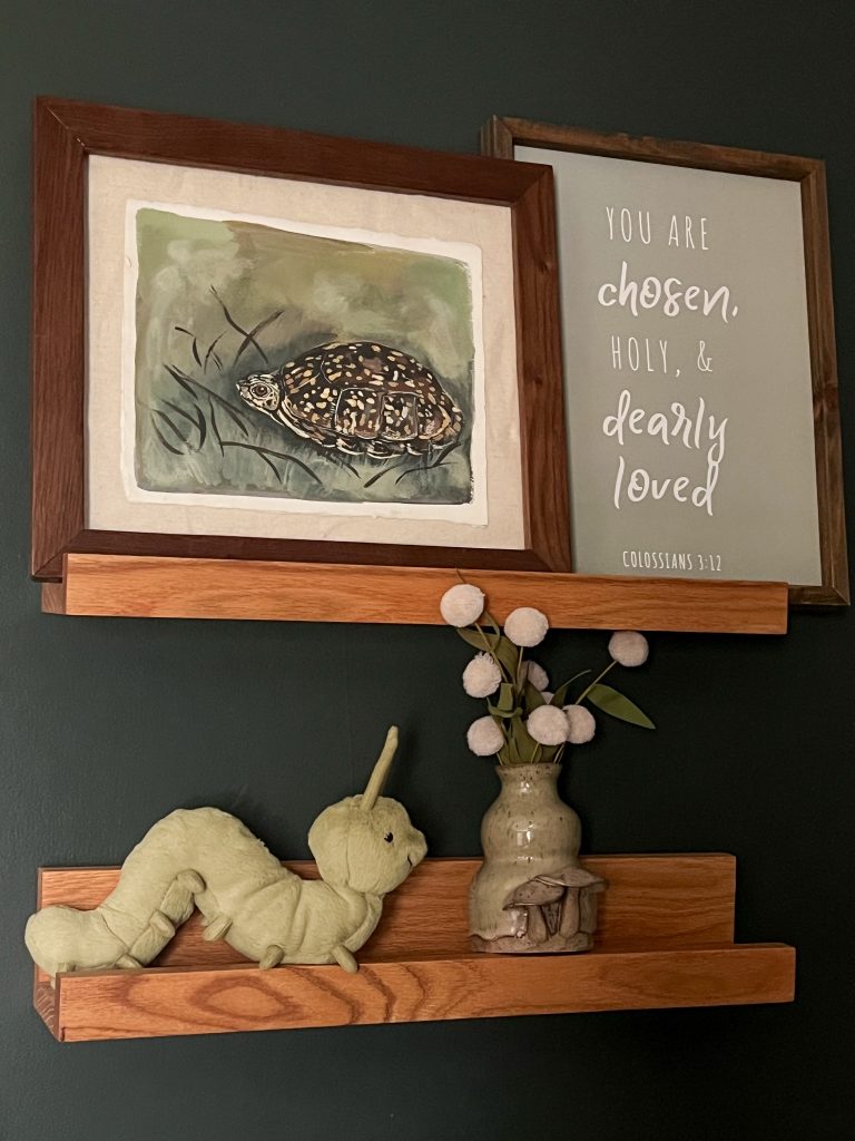 A cozy shelf display from The Midnight Mustard Seed featuring a framed art print of a spotted turtle on a green background with stylized grass, alongside a sign with the text “you are chosen, holy, & dearly loved” (Colossians 3:12) in elegant script. The wooden shelf, set against a dark green wall, also holds a plush caterpillar toy, a ceramic vase with dried white pom-pom flowers, and a small mushroom-topped figurine, creating a charming nature-inspired vignette.