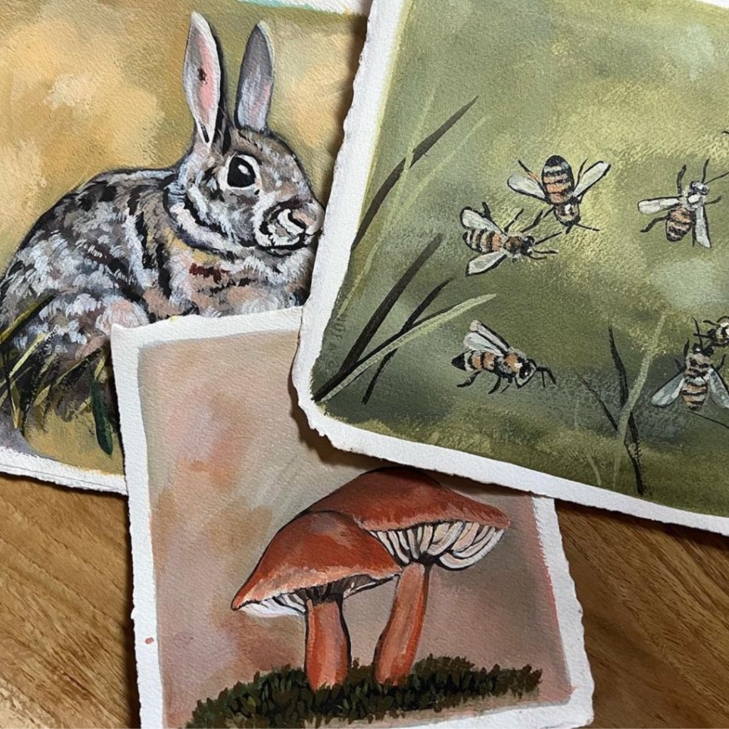 A collection of art prints from The Midnight Mustard Seed featuring nature-inspired illustrations. The prints include a detailed gray rabbit with pink ears on a yellow background, a group of bees flying over a green backdrop with stylized grass, and a pair of red-capped mushrooms with white gills on a soft peach base. The artworks are displayed on a wooden surface, showcasing earthy tones and delicate details.