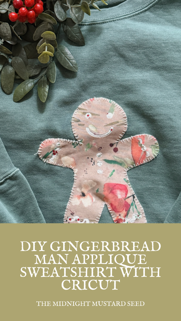 A close-up photo of a cozy green sweatshirt featuring a handmade gingerbread man appliqué in the center. The gingerbread man is cut from a light pinkish-beige fabric with a soft watercolor floral pattern, including red roses, green leaves, and small cherries. It has a smiling face with white icing-style eyes, red cheeks, a curved white smile, and small red candy-like buttons down its body. The edges are finished with a visible blanket stitch in white thread, giving it a rustic, handmade look. Surrounding the appliqué are scattered small red berries and green eucalyptus leaves on the sweatshirt, creating a festive, holiday-inspired vibe. The image is from a DIY craft tutorial titled “DIY Gingerbread Man Appliqué Sweatshirt with Cricut” by The Midnight Mustard Seed.