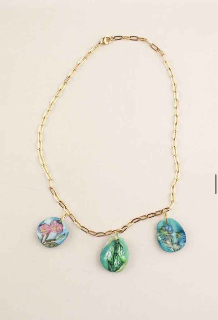 A product photograph of a handmade personalized ceramic multi-pendant birth flower necklace from The Midnight Mustard Seed, featuring three dangling ceramic charms on a delicate gold-colored figaro-style chain (elongated oval links) with a lobster claw clasp.
The three irregularly shaped, rounded ceramic pendants (teal/blue-green base glazes with subtle variations and textured, hand-painted details) hang at different lengths for a layered, asymmetrical look:
•  Left pendant: Round disc showing pink flowers (likely cosmos or similar) with yellow centers and green stems/leaves against a light blue sky-like background.
•  Center pendant (longest drop, teardrop/oval shape): Features delicate white lily-of-the-valley bells hanging from green stems and leaves, painted in soft greens on a turquoise/teal glazed background.
•  Right pendant: Another rounded disc depicting small blue/purple flowers (possibly forget-me-nots or irises) with green foliage on a similar teal/blue background.
The pendants have an artisanal, vintage-inspired aesthetic with watercolor-like glazing, subtle crackle or patina effects, and hand-carved/painted floral motifs that evoke personalized birth month symbolism (potentially representing months like January snowdrop/forget-me-not variant, May lily of the valley, and another spring/summer bloom). Displayed flat against a plain off-white/cream background, emphasizing the dainty, bohemian, nature-inspired jewelry style and colorful, nature-evoking details. By artists Samm Stafford of The Clay Moon and Midnight Mustard Seed and Whitney Winkler Art