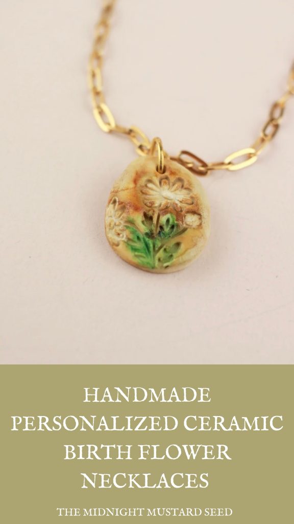 A close-up product photo of a handmade ceramic birth flower necklace from The Midnight Mustard Seed. The pendant is a small, irregularly shaped oval ceramic disc in warm earthy tones—mostly creamy beige with subtle orange-brown speckles and patina-like glazing. It features a delicately hand-carved and painted daisy-like flower with white petals and a yellow center, surrounded by smaller blooms and lush green leaves and stems at the bottom. The design has a vintage, textured, embossed appearance. The pendant hangs from a sturdy gold-colored chain with elongated oval links (figaro-style), attached via a small gold jump ring and bail. The overall style is dainty, bohemian, and personalized, displayed against a plain light background.