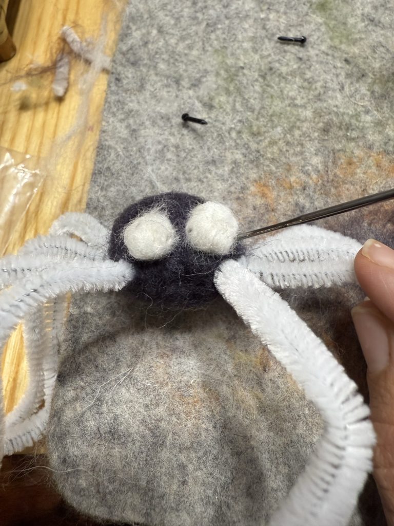 A close-up photo of a handmade needle-felted spider in progress on a fuzzy grayish-brown wool mat workspace. The spider’s round body is made from dark gray or charcoal wool felt, with two large, prominent white wool pom-pom-like eyes attached to the front. A long felting needle is actively being poked into the body area between the eyes, with the needle tip visible as it’s shaping or securing the features. The spider has eight fuzzy white pipe-cleaner legs extending outward in a radial pattern, giving it a cute, whimsical, slightly fuzzy appearance. Scattered around on the mat are small loose wool fibers, a few black bead-like pieces (possibly extra eyes or parts), and bits of crafting debris. The background shows a wooden surface and some crafting tools in soft focus. The overall scene captures a beginner-friendly needle felting project mid-creation, with warm indoor lighting highlighting the textured wool.