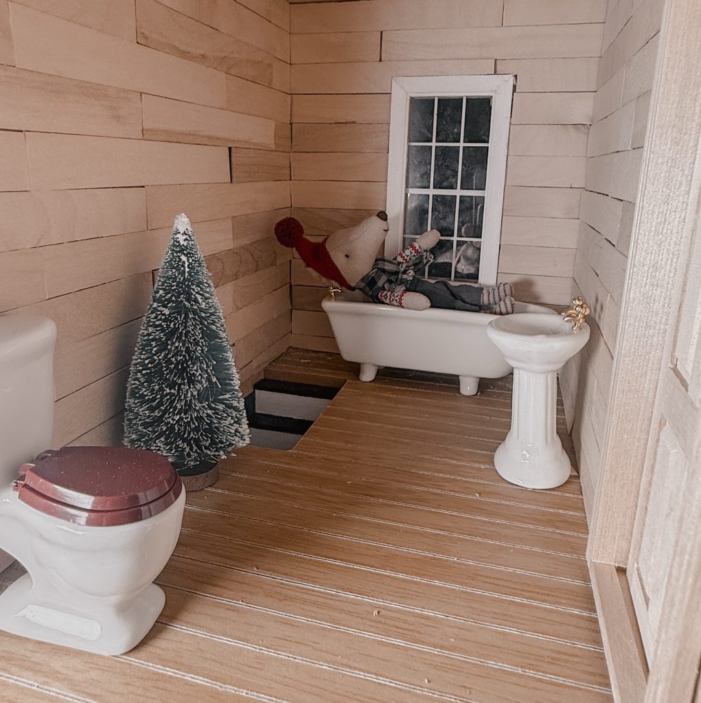 A cozy, whimsical miniature Christmas bathroom scene in a dollhouse crafted for Maileg mice, featured on my blog The Midnight Mustard Seed.
The small room features warm wooden plank walls and flooring, giving it a rustic cabin feel. A tiny white clawfoot bathtub sits in the corner, where an adorable felt Maileg mouse (wearing a red pom-pom hat, plaid sweater, gray pants, and striped red-and-white socks) relaxes luxuriously, leaning back with paws up and head resting on the tub edge, gazing out the white-framed window.
Next to the tub is a sweet pedestal sink with delicate gold faucets, while a classic white toilet with a red seat stands nearby. A small, snow-dusted miniature Christmas tree adds festive charm in the opposite corner, with tiny black steps leading up to the tub area.
This charming, holiday-inspired bathroom vignette is filled with warm, inviting details perfect for Maileg mouse collectors and dollhouse enthusiasts! 🎄🛁🐭
From The Midnight Mustard Seed blog.