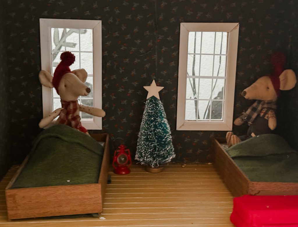 A cozy, enchanting miniature Christmas bedroom scene in a dollhouse designed for Maileg mice, featured on my blog The Midnight Mustard Seed.
The small room is warmly lit and wrapped in dark, rich wallpaper dotted with delicate red berries and evergreen sprigs, creating a festive yet intimate cabin-like atmosphere. Two white-framed windows let in soft light, framing the scene beautifully.
Two adorable felt Maileg mice are tucked into their beds for the night:
•  On the left, a mouse wearing a red pom-pom hat and a red-and-white plaid scarf snuggles under a lush green blanket in a wooden bed frame.
•  On the right, another mouse in a matching red pom-pom hat and cozy plaid shirt rests peacefully under a green blanket in an identical wooden bed.
Between the beds stands a tiny, snow-dusted Christmas tree topped with a sweet wooden star, its branches glistening with frost. A small red lantern with a glowing warm light sits at the base of the tree, adding a magical holiday glow to the room. A bright red wrapped gift rests on the wooden floor nearby, ready for Christmas morning.
This heartwarming bedtime vignette captures the quiet magic of the holiday season and is perfect for Maileg mouse lovers and dollhouse collectors! 🎄🛏️🐭
From The Midnight Mustard Seed blog. 🌱