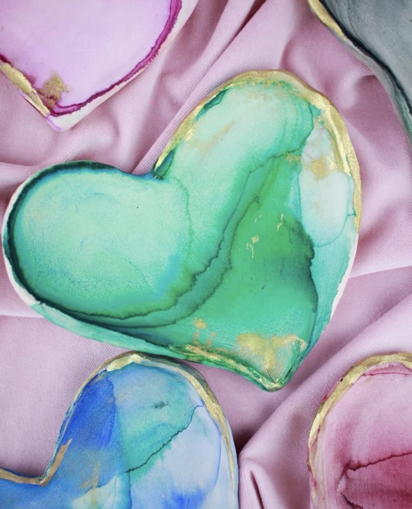 A collection of handmade heart-shaped ceramic trinket dishes displayed on soft pink satin fabric. Each dish features vibrant alcohol ink marbling with swirling, fluid patterns in pastel and jewel tones:
•  One in soft mint green and turquoise with deep emerald veins
•  One in rich blue fading to lighter shades
•  One in lavender pink with magenta accents
•  Others in mixed blues, greens, and purples
All dishes have a white base, irregular organic edges, and are elegantly finished with gold leaf along the borders, adding a luxurious metallic shimmer. The pieces are arranged overlapping slightly, showcasing their glossy, translucent surfaces and unique fluid art designs.
Image from the blog: www.themidnightmustardseed.com