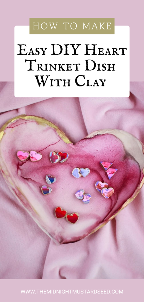 A promotional graphic for a DIY tutorial featuring a close-up of a handmade heart-shaped ceramic trinket dish resting on soft pink satin fabric. The dish showcases a beautiful alcohol ink marbled design with swirling, fluid patterns in soft pink, lavender, and white tones, accented by delicate gold leaf edging for a luxurious finish.
Inside the glossy, shallow heart-shaped dish are scattered several small, colorful heart-shaped enamel pins in shades of pink, red, blue, and iridescent purple.
The image includes overlaid text in elegant styling:
•  “HOW TO MAKE” in a gold-framed box
•  “EASY DIY HEART TRINKET DISH WITH CLAY”
•  Website URL: www.themidnightmustardseed.com
The overall color palette is romantic and pastel, with a feminine, dreamy aesthetic.